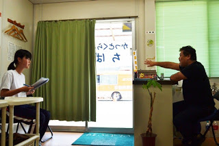 プレハブのような室内で、女子生徒が椅子に座ってノートを持ちながら、カウンター越しに男性の話を聞いている様子が写っている写真