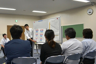 ホワイトボードの横に立って資料を説明する男性と、それを椅子に座って見ている参加者4人の後ろ姿の写真