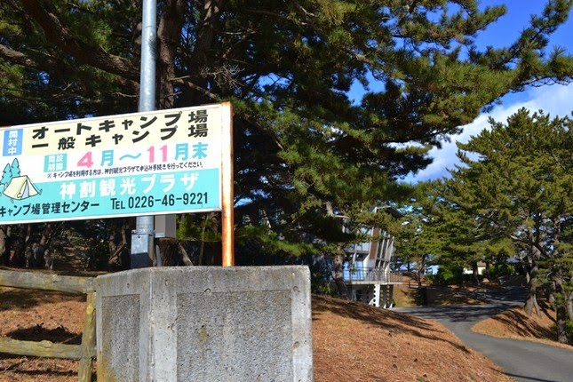 「オートキャンプ場・一般キャンプ場」と書かれた案内板のそばに松林が広がり、奥にはコテージと舗装された道が見える様子を写した写真