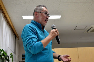 青いフリースを着用した男性がマイクを手に何かを熱心に話している様子を横から写した写真