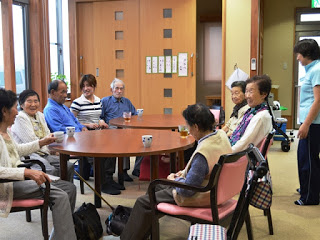 自然光が差し込む窓のある室内で、2つ並べて置かれた丸いテーブルの周りの椅子に高齢の男女7人と一人の女性が座り、和やかに会話をしている様子の写真