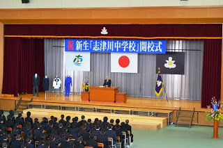 国旗と町旗、校旗と「祝 新生「志津川中学校」開校式」と書かれた横断幕が掲げられたステージ左側に制服がかかった4体のマネキンと、右側に旗が設置され、中央の檀上に立つ男性の話を、生徒たちが聞いている様子を2階から撮影した写真
