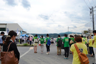 黄色や緑のTシャツを着た多くの人々が屋外に集まり、黙祷を行っている様子を後方から撮影した写真