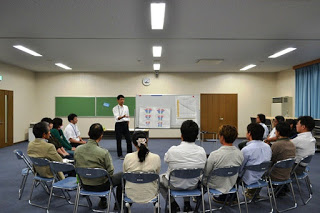 教室の中で半円を描いて並んだ椅子に座った参加者たちが、前方のホワイトボード横に立っている男性の話を聞いている様子の写真