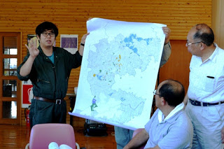 1人の人物が大きな地図を広げて立ち、横に立った男性が説明をしている様子を2人の男性が地図を見ながら聞いている様子の写真