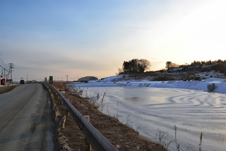 雪に覆われた冬の風景の中、凍りかけた川と並行して走る道路が静けさを漂わせ、朝焼けの光が景色を柔らかく照らす冬の川辺の写真