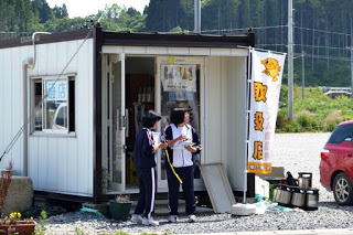 商品が陳列されている小さな建物の前で、ジャージ姿の2人の女子生徒がメモを取りながら話し合っている様子が写っている写真