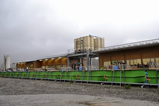 緑色の長い水槽が横に並び、その後ろに屋根の上にタンクを乗せた木造の建物が見える、水産物を扱う加工施設の外観全体の写真