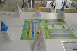 教室の机に「みんなのまちづくりゲーム」のボードと、青・緑・黄・赤などの色のピラミッド型の駒が円を描くように配置されている写真