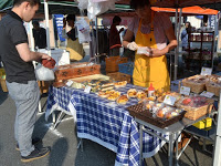 屋外の市場で青と白のチェック柄のテーブルクロスがかけられたテーブルの上に、パンや焼き菓子などが種類豊富に並べられ、黄色のエプロンを着た店員の男性が男性客の接客をしている様子の写真