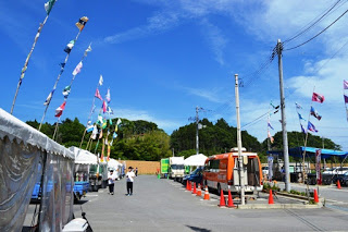 晴れた空の下、色とりどりの旗が風に揺れ、仮設テントやキッチンカーが並ぶ広場に数人の生徒が歩いている様子が写っている写真