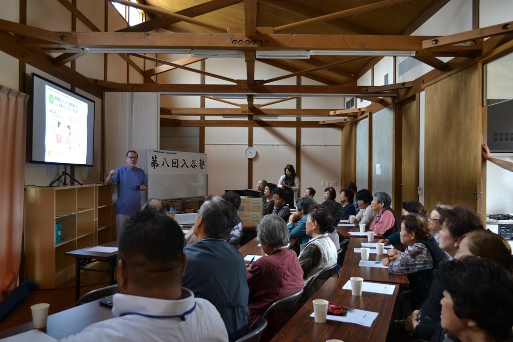 木造の部屋で多くの人が机に座って前方のスクリーンを見ながら講義を受けている様子を写した写真
