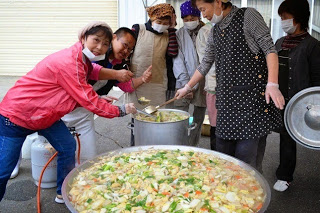 大鍋にたっぷりの野菜が入った豚汁を、笑顔の女性たちが周囲でかき混ぜたり、カメラに向かってポーズをとったりしている様子の写真