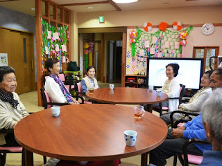 大きなテレビの前で、湯飲みが置かれた二つの丸い木製テーブルを囲んで人々が座り、会話を楽しんでいる様子の写真