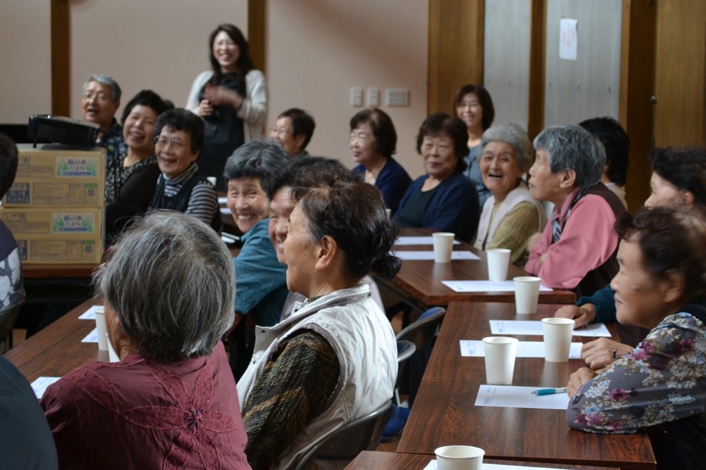 飲み物のカップが置かれた長机の席に座った大勢の参加者たちが、笑顔で楽しく講義を受けている写真