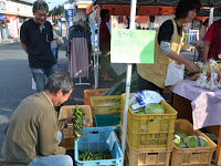 青空の下で開かれた市場において、テーブルの上に並べられた新鮮な野菜を売るために人々が立ち寄っている様子が写っており、にぎやかで地域の交流が感じられる青空市場の写真