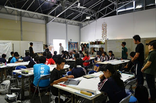 机には資料や文房具が置かれている複数の白いテーブルが設置された室内で、生徒たちが数人の教師に見守られながら、黒板の前に立つ発表者の話を聞いている様子が写っている写真