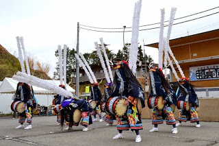 色鮮やかな衣装を着て、体の前に大きな太鼓を持ち、頭に白い紙飾りを高く立てた踊り手たちが一斉に舞を披露している様子の写真