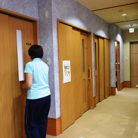 明るい紫色の壁に囲まれた廊下の片側に複数の木の扉が並び、そのうちの一つの前に女性が立っているところを後方から写した写真