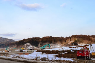 背景に木々に覆われた丘が広がり、雪が積もる中、重機やトラックが点在する冬の工事現場の写真