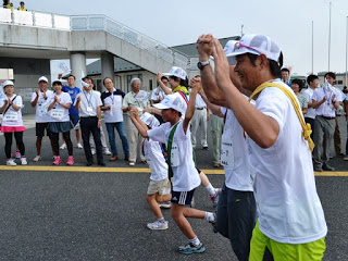 タスキをかけた数人の大人と子供が手を繋いで楽しそうに走っている様子を、沿道の人々が応援している写真