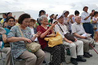 屋外で並べられた椅子に沢山のお客さんが座り、拍手をしながら舞台の方を楽しそうに見つめている様子が写っている写真
