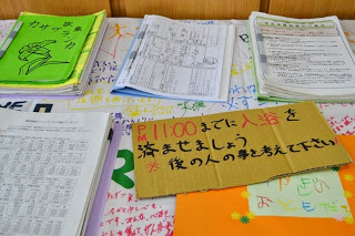 東日本大震災の資料やノート、カラフルなメモがテーブルに広げられていて、中央に「午後11:00までに入浴を済ませましょう」と書かれた段ボールの掲示がある資料が展示されている様子を写した写真