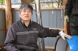 屋根付き作業場の中で、青いドラム缶の装置を前に手を添えながら説明している炭焼き指導者の阿部さんをアップで撮影した写真