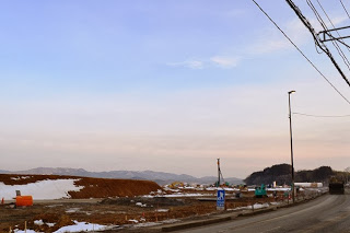 道路が整備された左側に、雪が残る地面に土砂や資材が広がり、重機が点在する工事現場の奥に山々がそびえる風景の写真