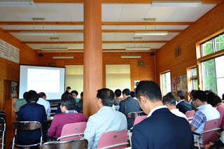 木の柱と大きな窓がある部屋で、複数の人々が椅子に座ってスクリーンの方を向いており、セミナーに参加している様子が写っている写真