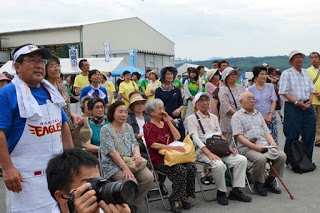 椅子に座った高齢者や立っている人たちが、正面のステージを見つめながら楽しそうに笑顔を見せている写真