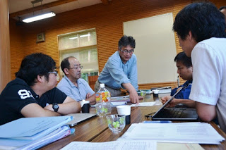 5人の男性がテーブルを囲んで座り、1人が立ってテーブルに広げた資料を指し示している様子を写した写真