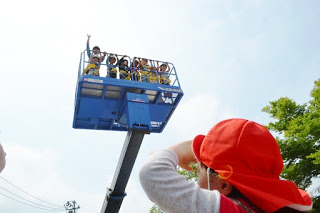 青い高所作業車のバケットに乗って高所に上った園児たちが手を振っており、それを地上から赤い帽子をかぶった園児が見上げている様子の写真