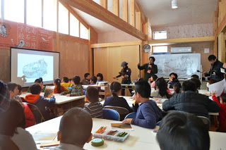 教室のような空間で、スクリーンに映し出された化石の画像を見ながら講師の話を聞いている多くの子どもたちの写真