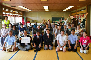 畳の部屋で正装した地域の関係者や参加者たちが、カメラに向かって笑顔で集合写真を撮っている様子が写っている写真