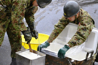 ヘルメットを被り迷彩服を着た自衛隊員が、炊き出しで炊きあがった大型の釜から取り出したご飯を、大きなビニール袋をしいたクーラーボックスの様な容器に移し替えようとしている様子の写真