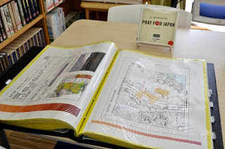 テーブルの上に東日本大震災の『復興見える化』のプリントアウトされた冊子が開かれて置かれていて、テーブルの奥には『PRAY FOR JAPAN』と書かれた小さな立て看板がある展示の様子を写した写真