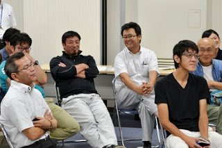 パイプ椅子に座った男性たちが、笑顔で前を向いて話を聞いている様子の写真