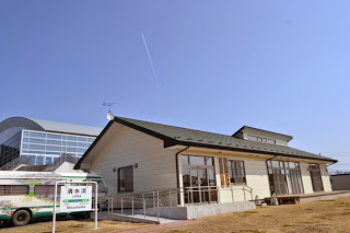 青空の下に建つ平屋の建物の右側には大きな窓が並び、左側にはスロープ付きの入り口があり、建物の左手前には「青水道」と書かれた看板と、同じ名前のバスが停まっている様子の写真