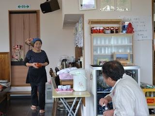 黒いエプロンをした女性が、厨房からテーブルに向かってお椀を運んでいる最中で、手前には男性が椅子に座って振り返っている様子の写真