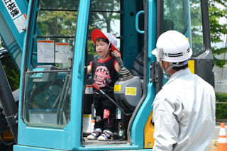 帽子をかぶった男の子がショベルカーの操縦席に立ってレバーに手をかけながら楽しそうにしており、下から白いヘルメットをかぶった作業員が安全を見守っている写真