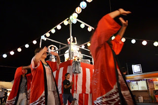 夜の祭りの会場で、赤い法被を着た人々がやぐらの周りで輪になって踊っている様子を写した、活気ある盆踊りの写真