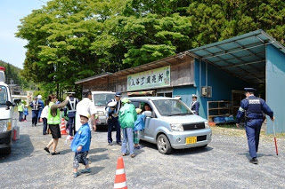 制服を着た警察官数人と青いスモッグを着た園児たちが、入谷サン直売所前と書かれた建物の前で、停まった車両の運転手や同乗者とやり取りをしている様子の写真