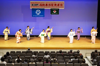 舞台の上で色とりどりの着物を着た出演者たちが優雅に踊りを披露し、客席の人々が見守る発表会の写真