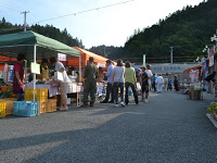 緑豊かな山々を背景に設けられた青空市場に緑やオレンジのテントが並び、買い物客でにぎわっている様子の写真