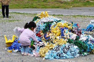 地面に山のように積み上げられたたくさんの千羽鶴の山に、ピンクの上着を着た小さな女の子が千羽鶴を並べている様子の写真