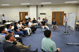 会議室の中で、ホワイトボードの前に半円型に並べた椅子に座った参加者たちが、ホワイトボードの横に立って説明をする男性の話を聞いている様子の写真