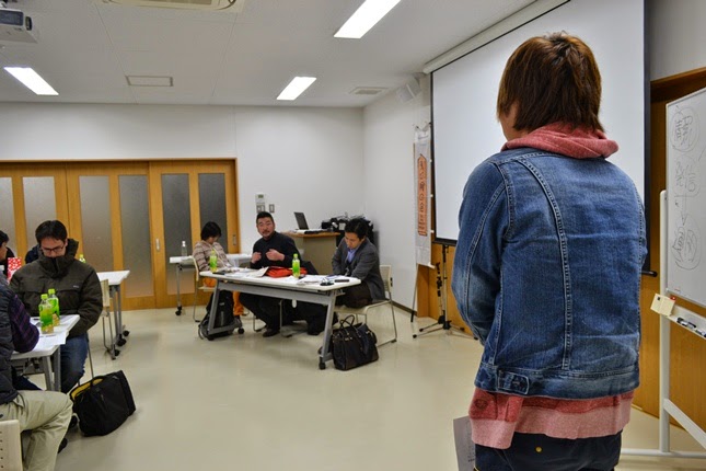 ジージャンを着た人物がスクリーンの前に立って発表しており、他の参加者が自席に座って聞いている写真