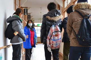 木造校舎のような建物の廊下で、ガイドの女性がマスクをして訪問者に説明をしており、来訪者たちはリュックを背負いながら話を聞いている案内中の写真