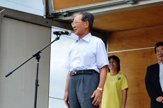 屋外ステージで年配の男性がマイクの前に立ち、観客に向かってあいさつをしている様子の写真
